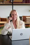 Vrouw met blond haar achter haar laptop met telefoon aan haar oor